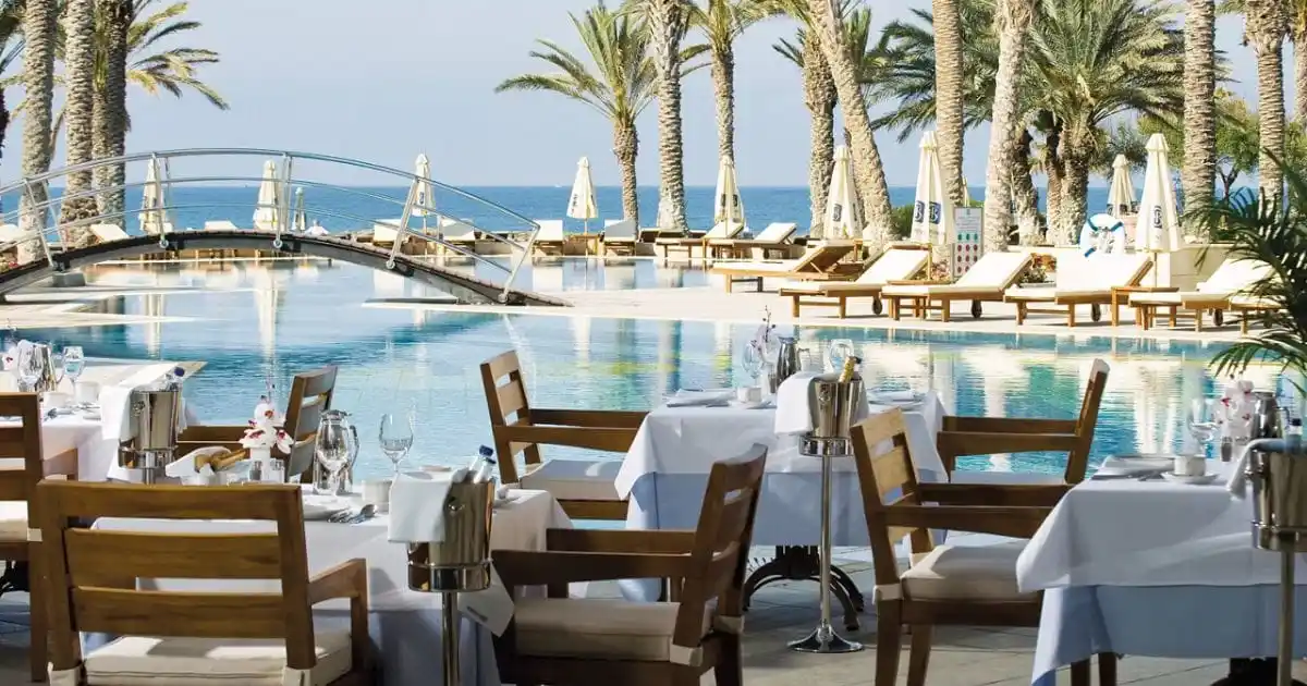 urant tables set with white tableclothes next to the swimming pool, an arched footbridge, and palm trees at the Asimina Suites Hotel in Paphos, Cyprus.
