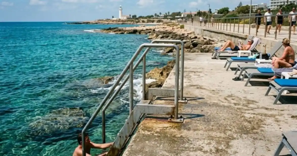 A paved concrete sunbathing platform with a ladder leading into the sea, and a lighthouse in the background along the Kato Pahos coastal path in Cyprus.