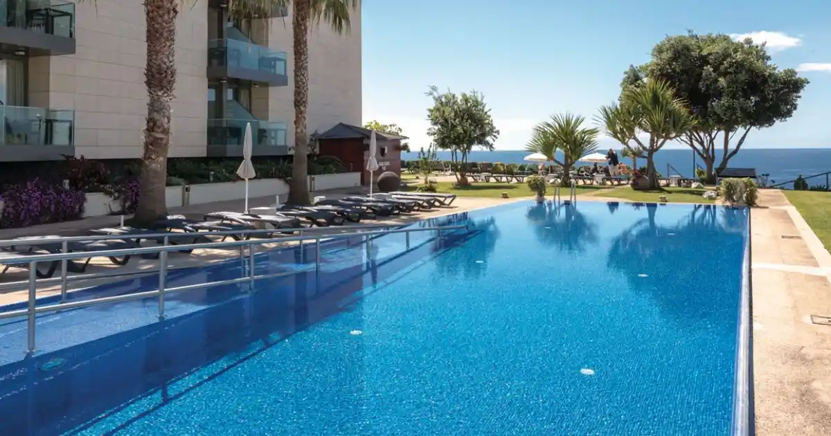 The Golden Residence hotel outdoor pool and sounloungers backe by palm trees and an view of the Atlantic ocean.