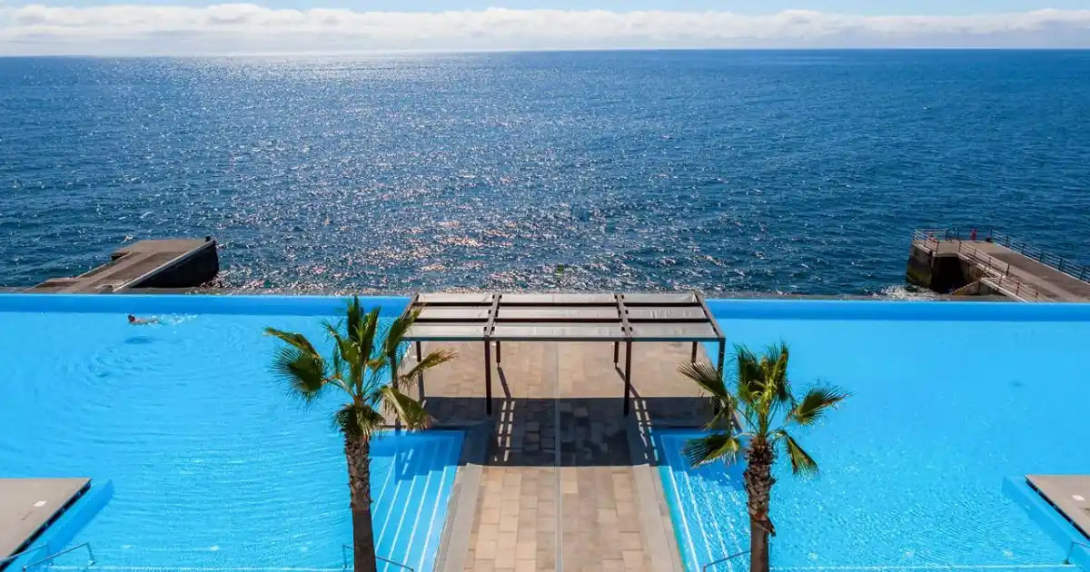 View of the infinity pool with deck and palm trees, at the VidaMar Resort Madeira, Funchal, overlooking the Atlantic ocean.